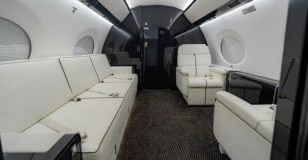 The business jet cabin interior featuring a white leather sofa, individual seats, and dark patterned carpet.