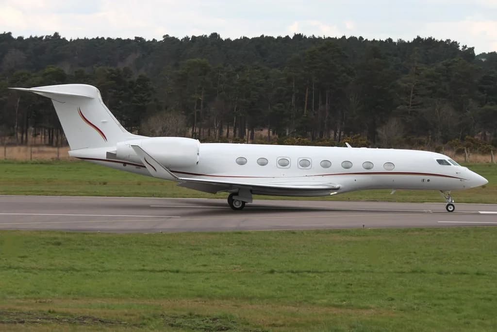 A white Gulfstream business jet with red and gold stripes on the tail and fuselage, on a runway with grass and trees in the background.
