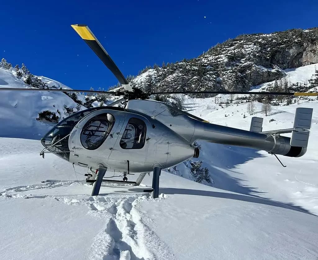 A silver helicopter with yellow-tipped rotor blades is landed in deep snow on a sunny mountain slope.