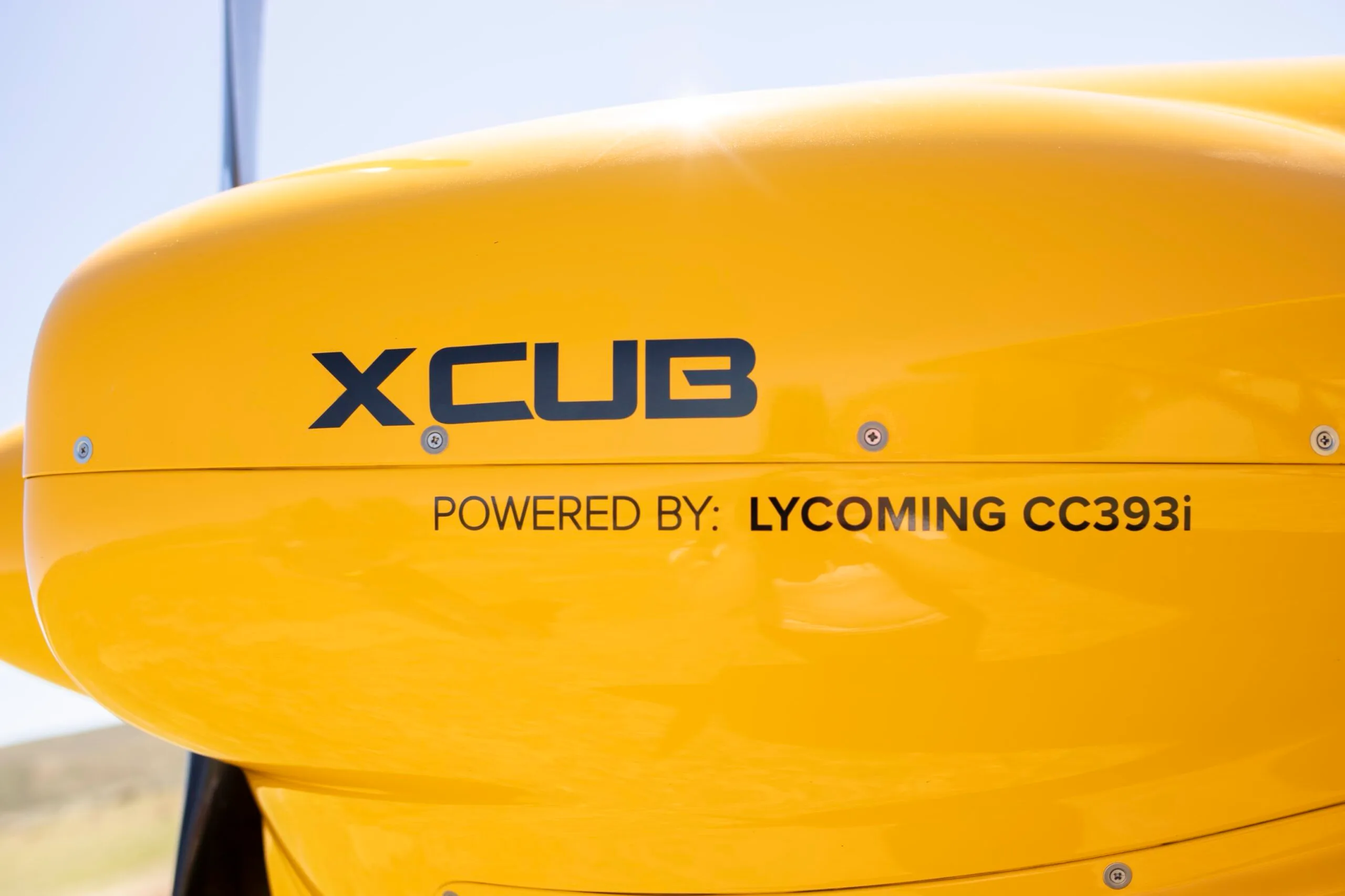 A close-up of the yellow engine cowling of the XCub, showing "XCUB" and "POWERED BY: LYCOMING CC393i" text.