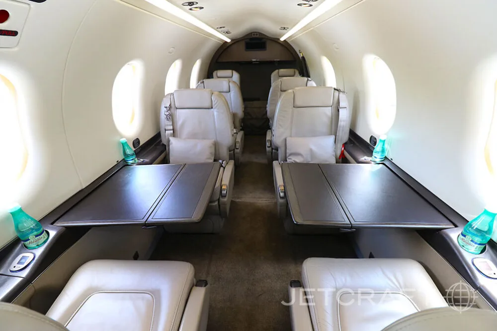 The interior cabin of a Pilatus PC-12 aircraft showing light beige leather seats and two fold-out tables.