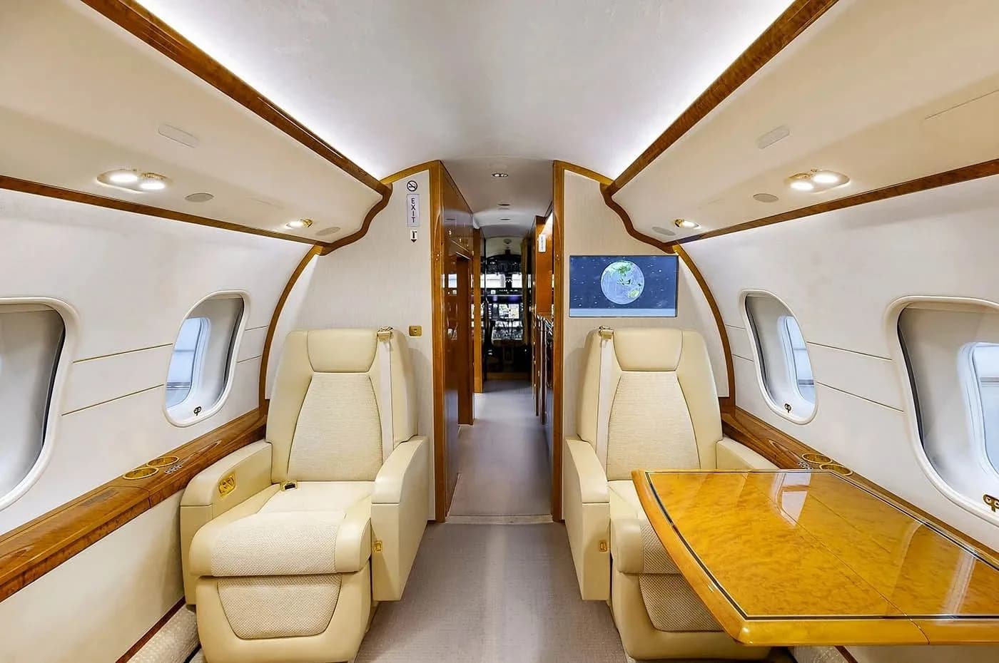 A luxurious aircraft cabin interior with two cream-colored reclining seats, a wooden table, and a screen displaying a globe.