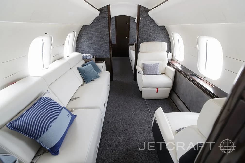 A private jet cabin featuring a white sofa with blue pillows and a single white armchair.