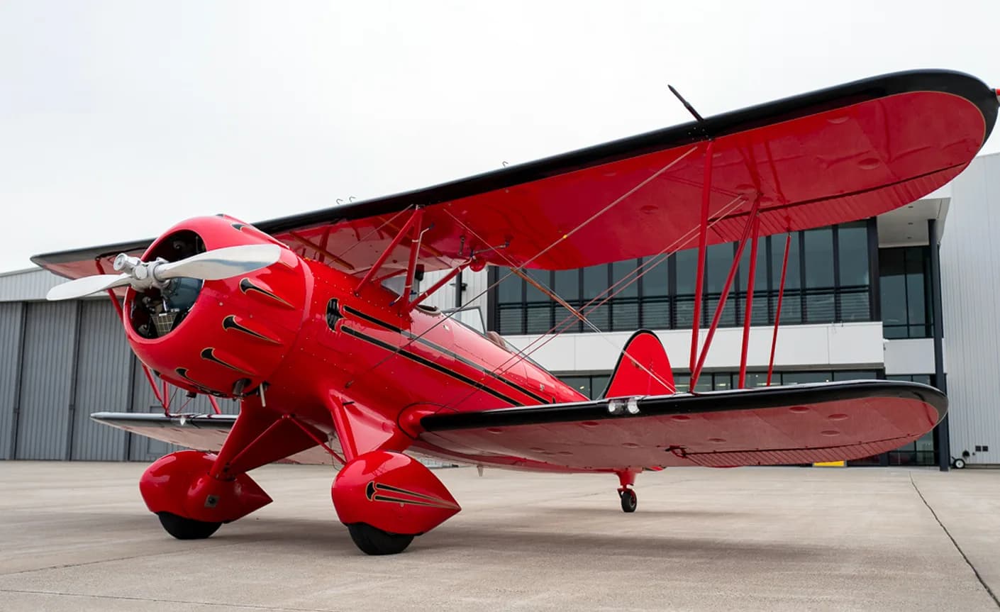 A red biplane sits on the tarmac in front of a modern building.