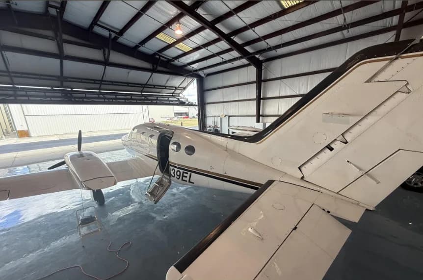 A rear-left view of the white Cessna 414 aircraft with registration N239EL, parked in a hangar with its door open and steps down.