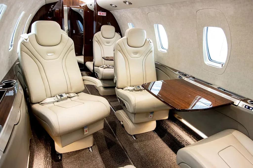 A close-up view of the luxurious interior cabin of a private jet shows light tan leather seats with a wooden pull-out table.
