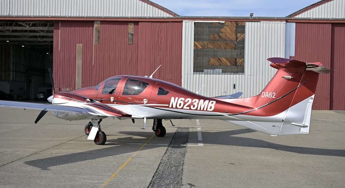A maroon and silver Diamond DA62 aircraft is parked on the tarmac in front of a hangar.
