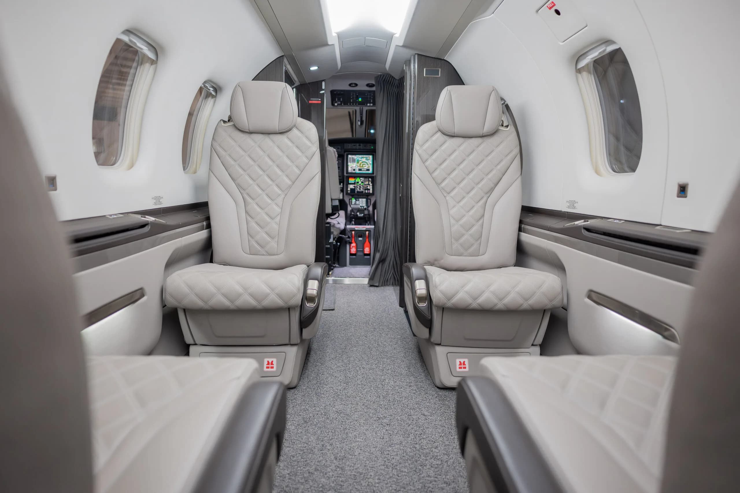 The aircraft cabin shows light grey quilted leather seats looking forward towards the cockpit, which is partially visible.