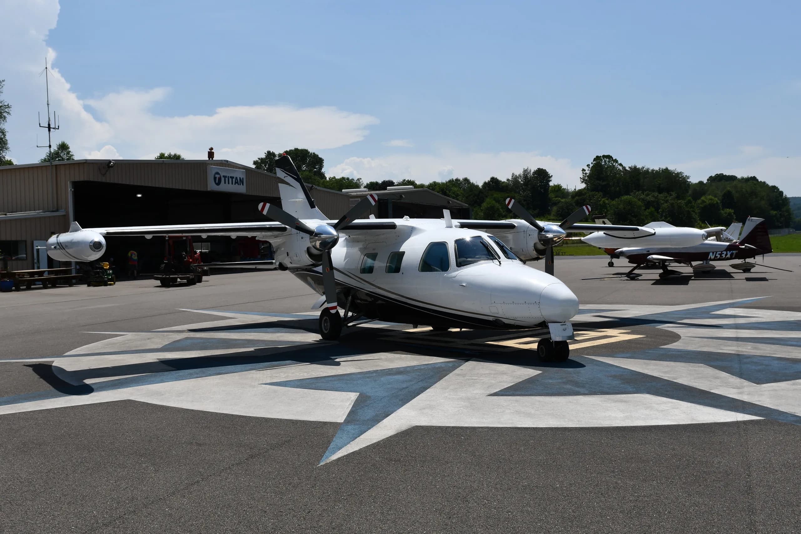 A white twin-engine propeller aircraft with dark stripes is parked on a compass rose at an airfield, with a hangar and trees in the background.
