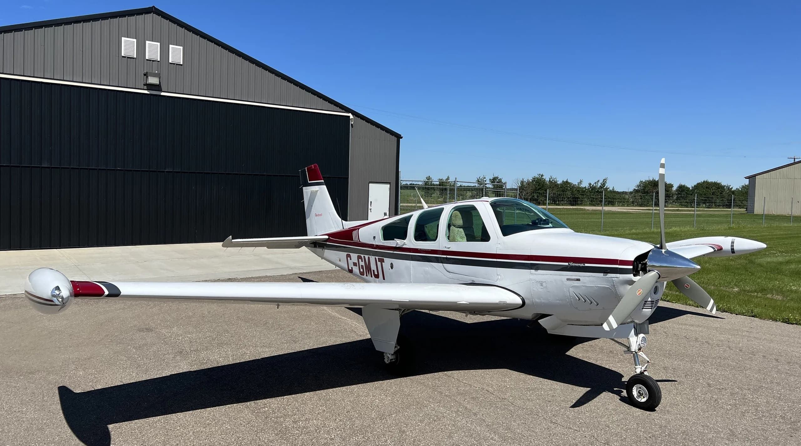 A white Beechcraft Bonanza with maroon and black stripes, registration C-GMJT, is parked on an airfield in front of a hangar under a clear blue sky.