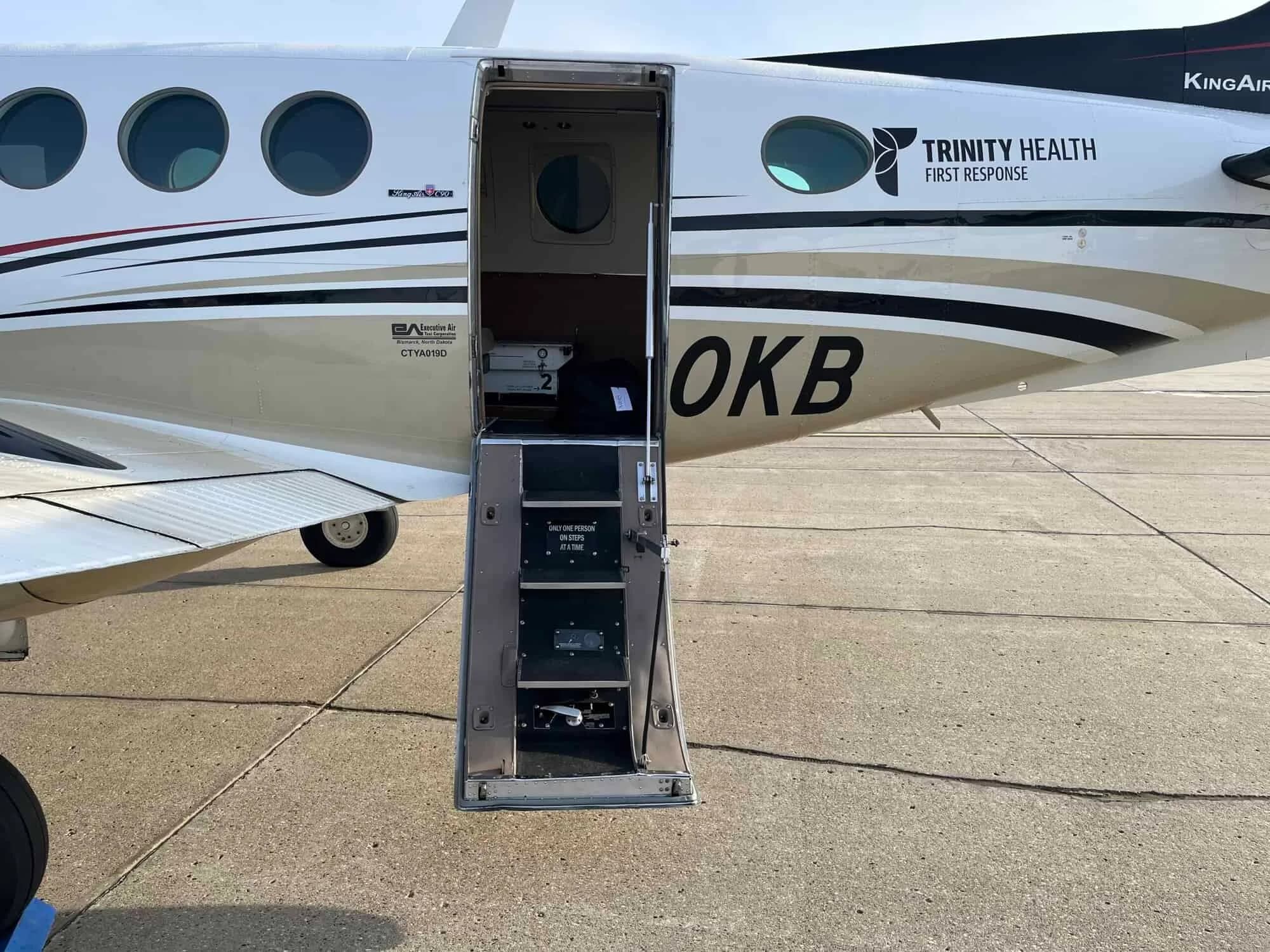 The open airstair door of a white and gold aircraft with "TRINITY HEALTH FIRST RESPONSE" text on the fuselage.