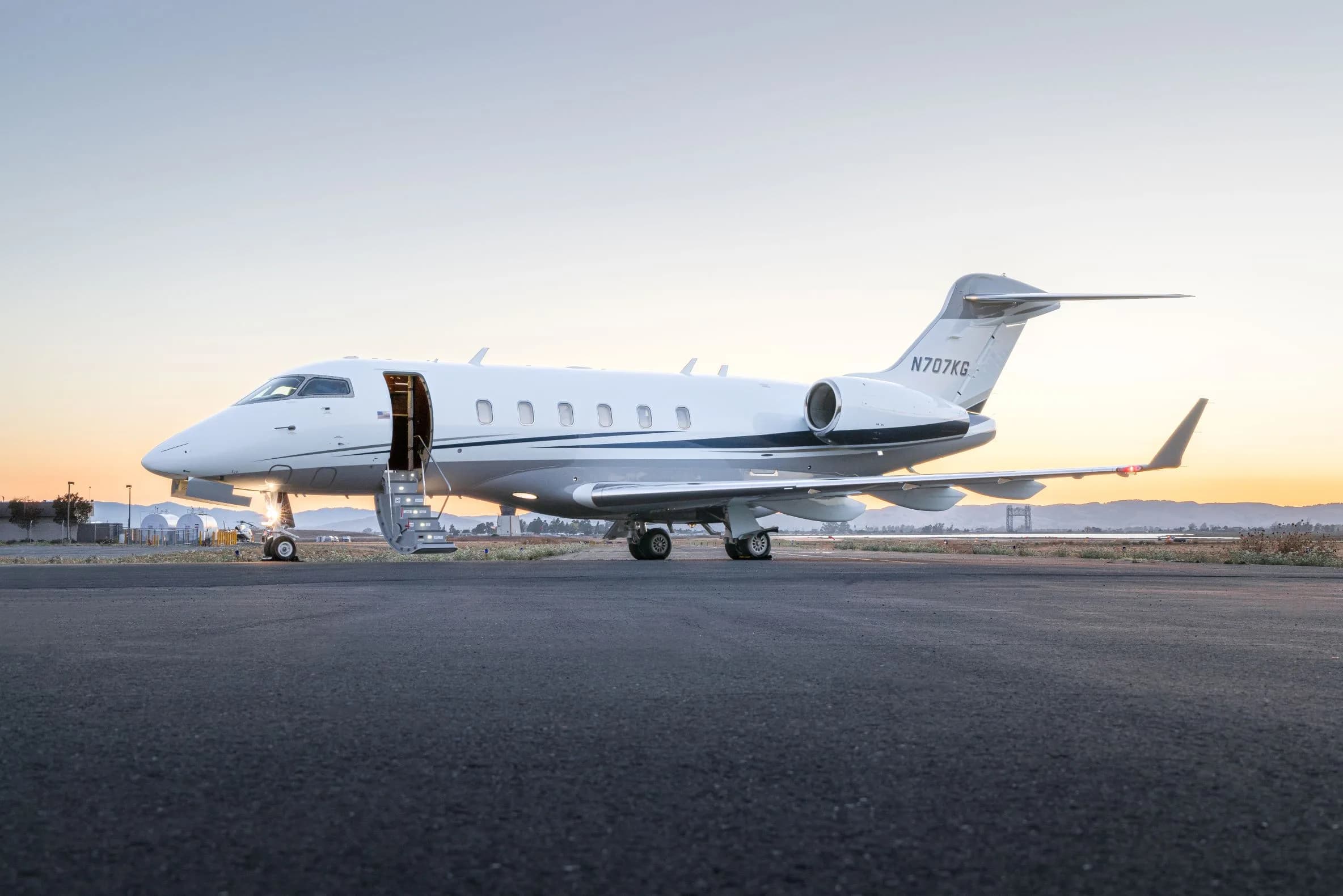 A white and grey business jet with its door open and stairs extended, parked on an tarmac at sunset.