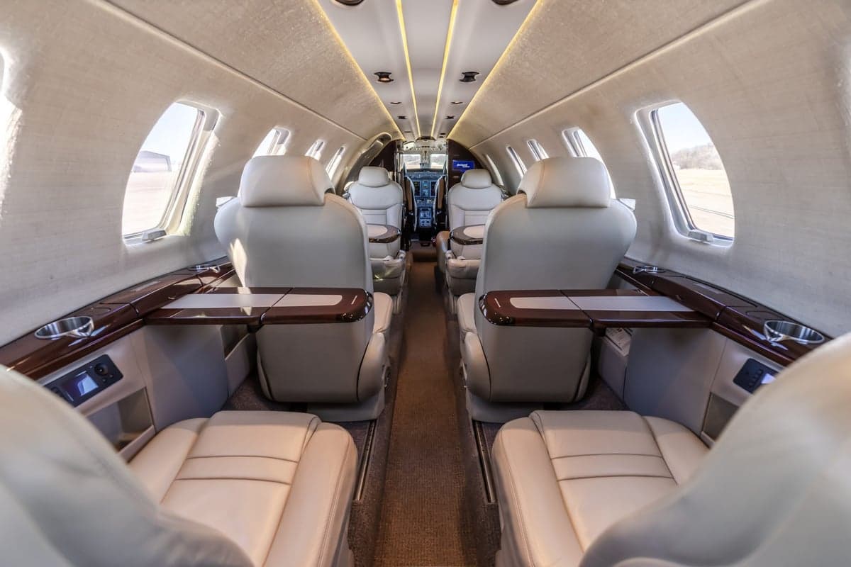 The luxurious interior cabin of a private jet features light beige leather seats, wooden tables, and an aisle leading to the cockpit.