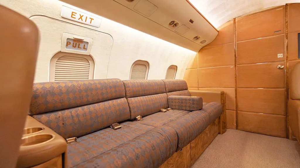 A close-up of the private jet cabin's patterned sofa with seatbelts and an "EXIT" sign above a window.