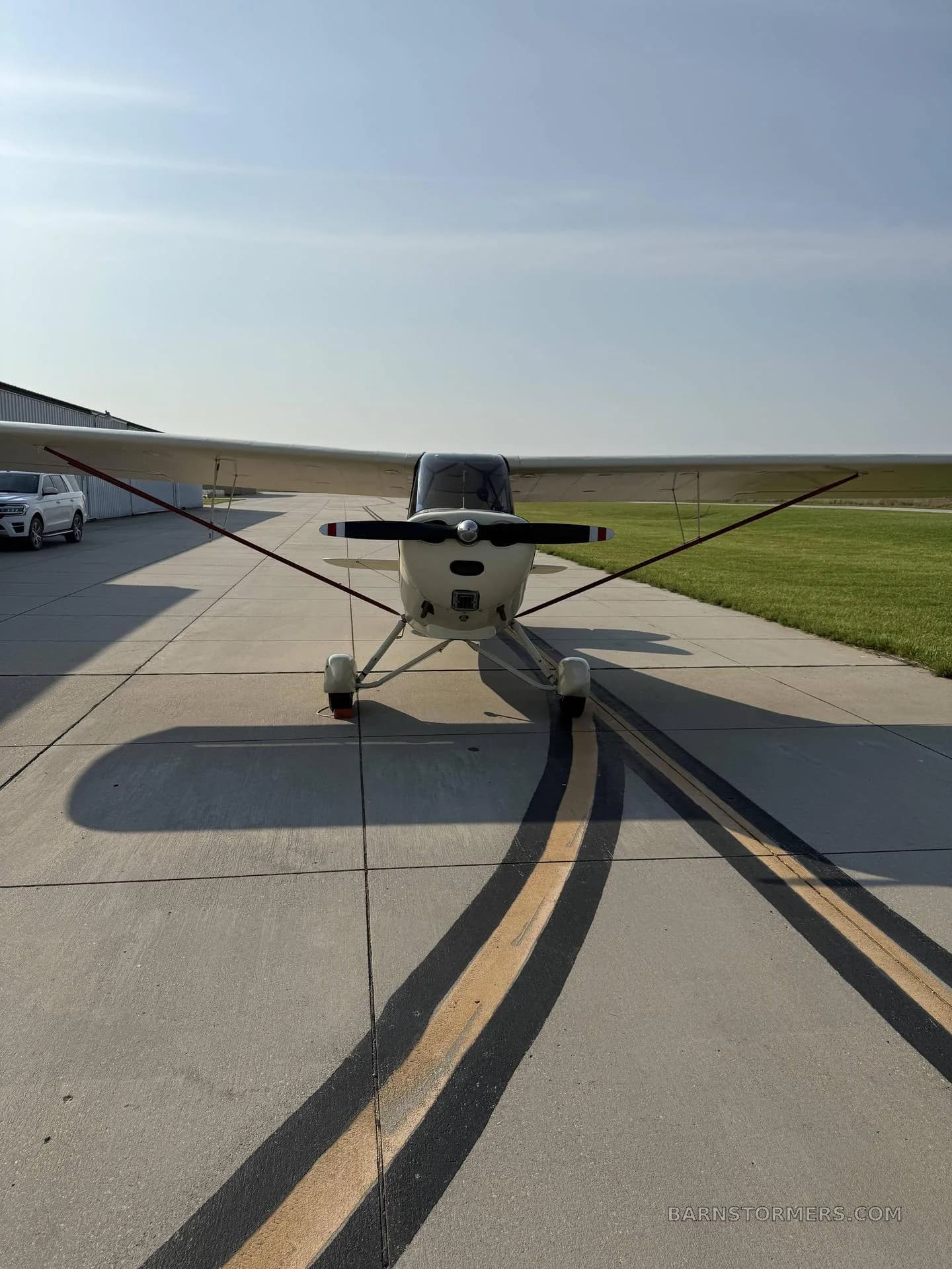 A cream-colored, high-wing taildragger aircraft with a black propeller and red stripes, viewed head-on on a concrete tarmac.