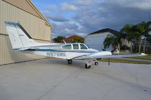 A Beechcraft Bonanza is parked on the tarmac with palm trees in the background.