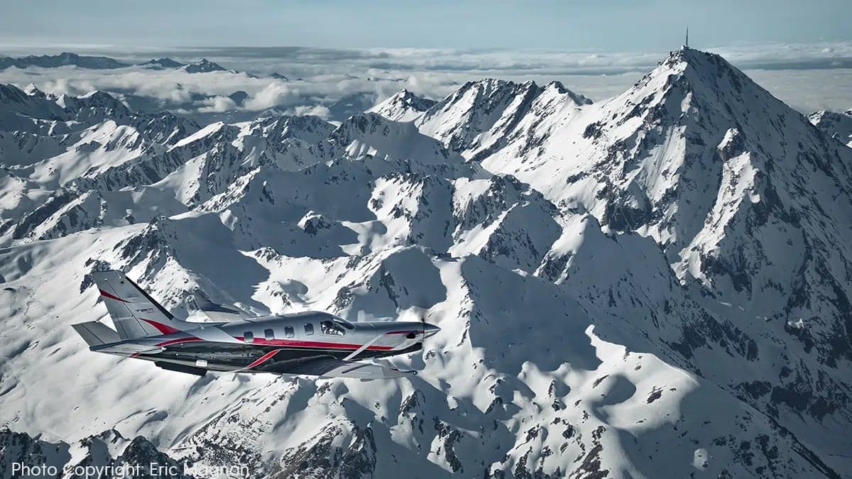 A TBM 960 aircraft with a silver, black, and red paint scheme flying over snow-capped mountains.