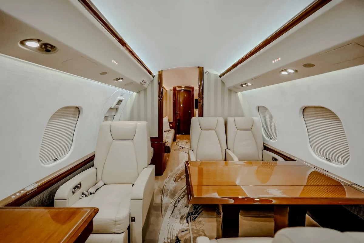 A private jet cabin interior with light-colored seats, wooden tables, and a view towards the rear compartment.
