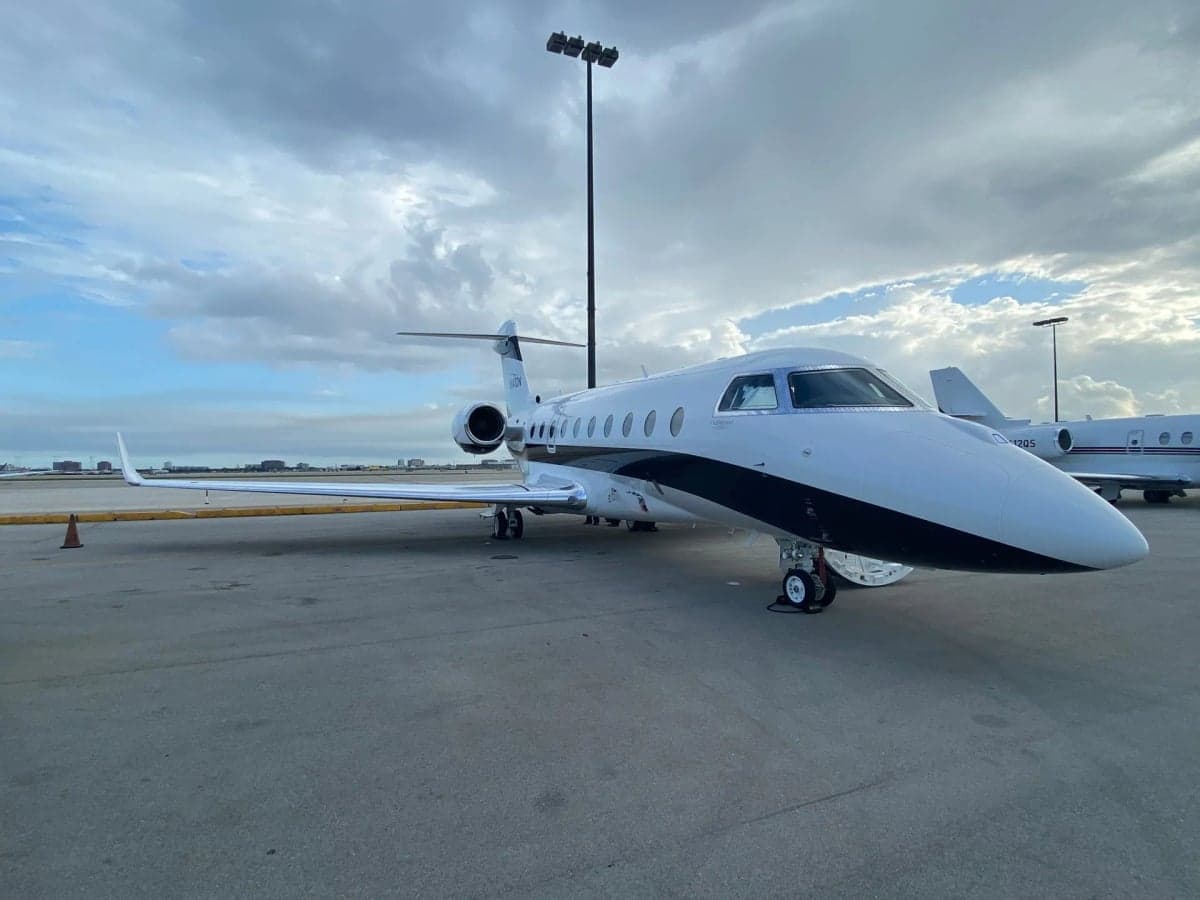 A white business jet with black and grey stripes, parked on an airport tarmac under a dramatic cloudy sky with another aircraft nearby.
