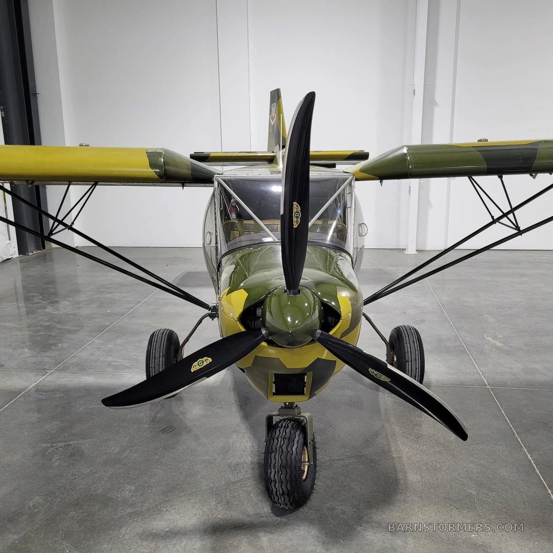 A head-on view of a camouflage-painted, high-wing aircraft with a large propeller, parked inside a hangar.