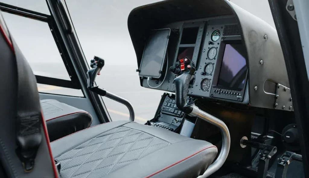 The cockpit of a helicopter featuring multiple screens, flight controls, and a grey quilted pilot seat.