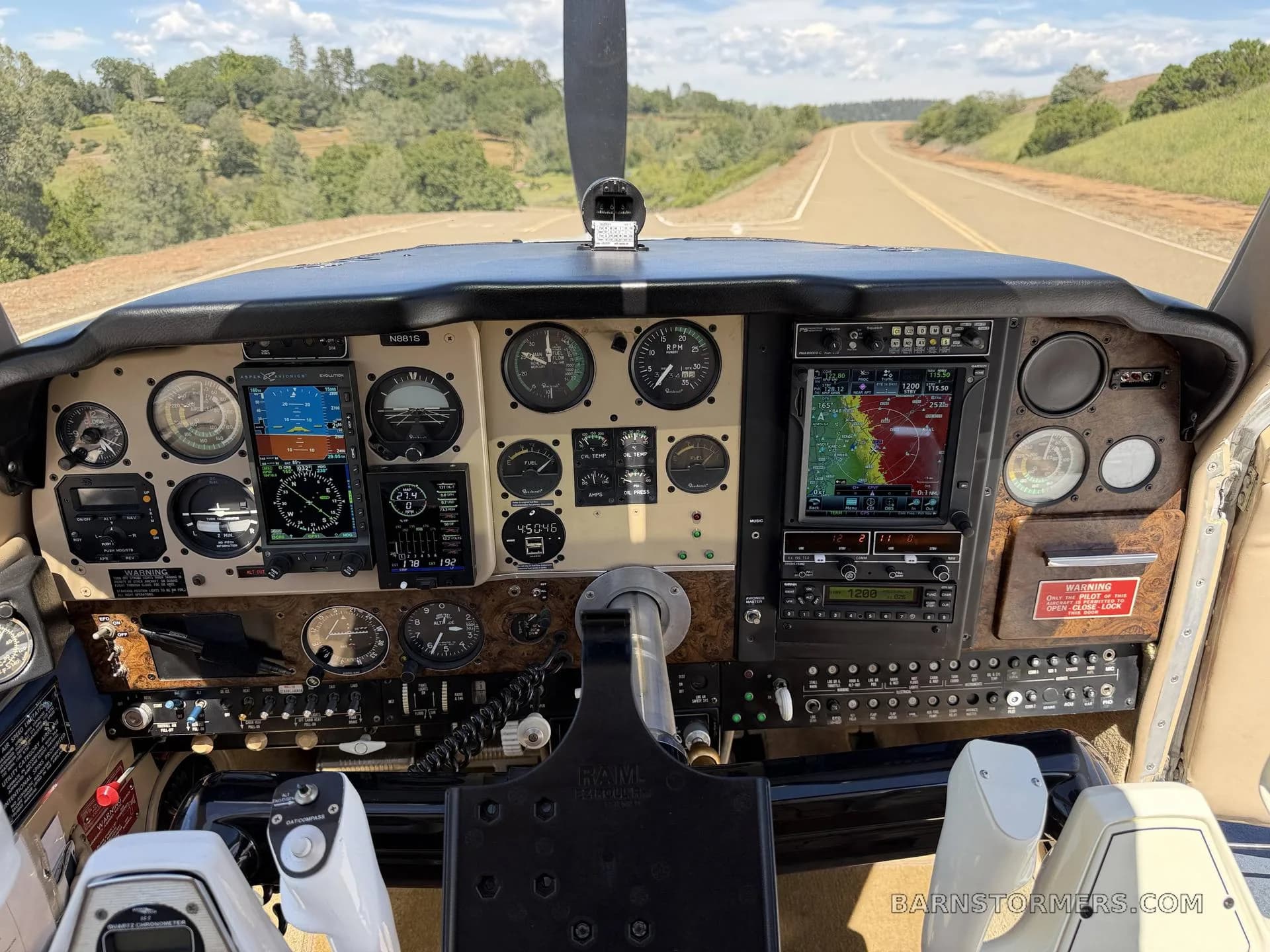 The cockpit of a Beechcraft Bonanza showing the instrument panel with multiple screens and gauges, looking out onto a road.