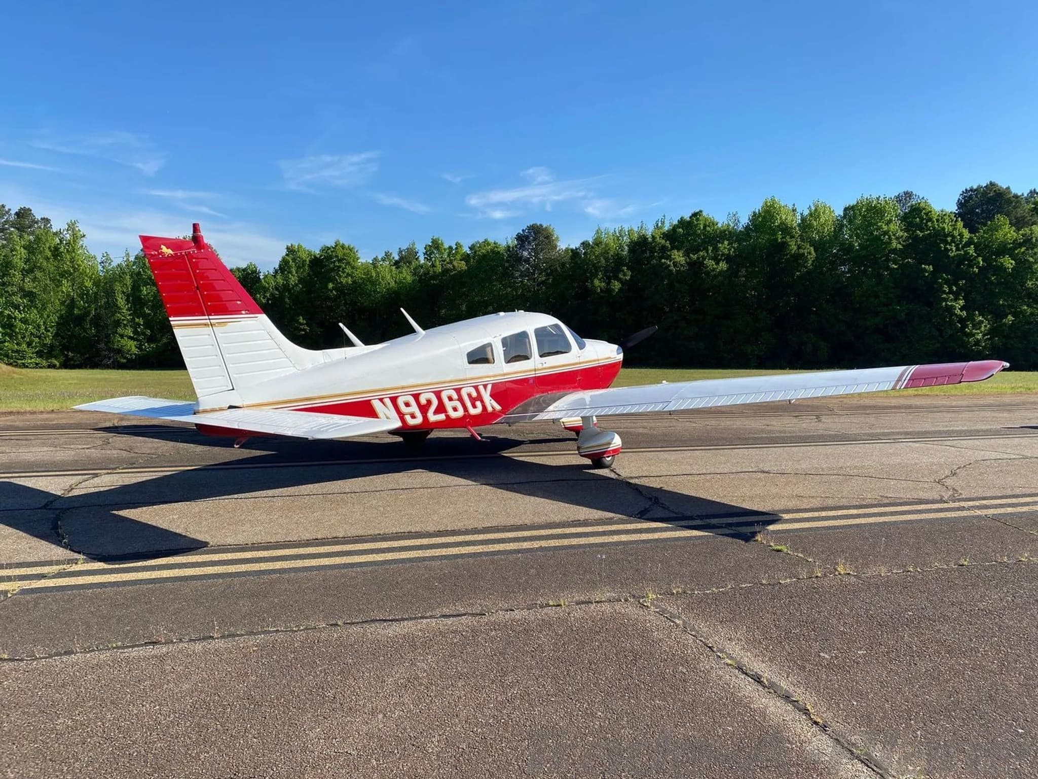 A side view of the white and red Piper Arrow II (N926CK) parked on an asphalt runway with trees in the background.