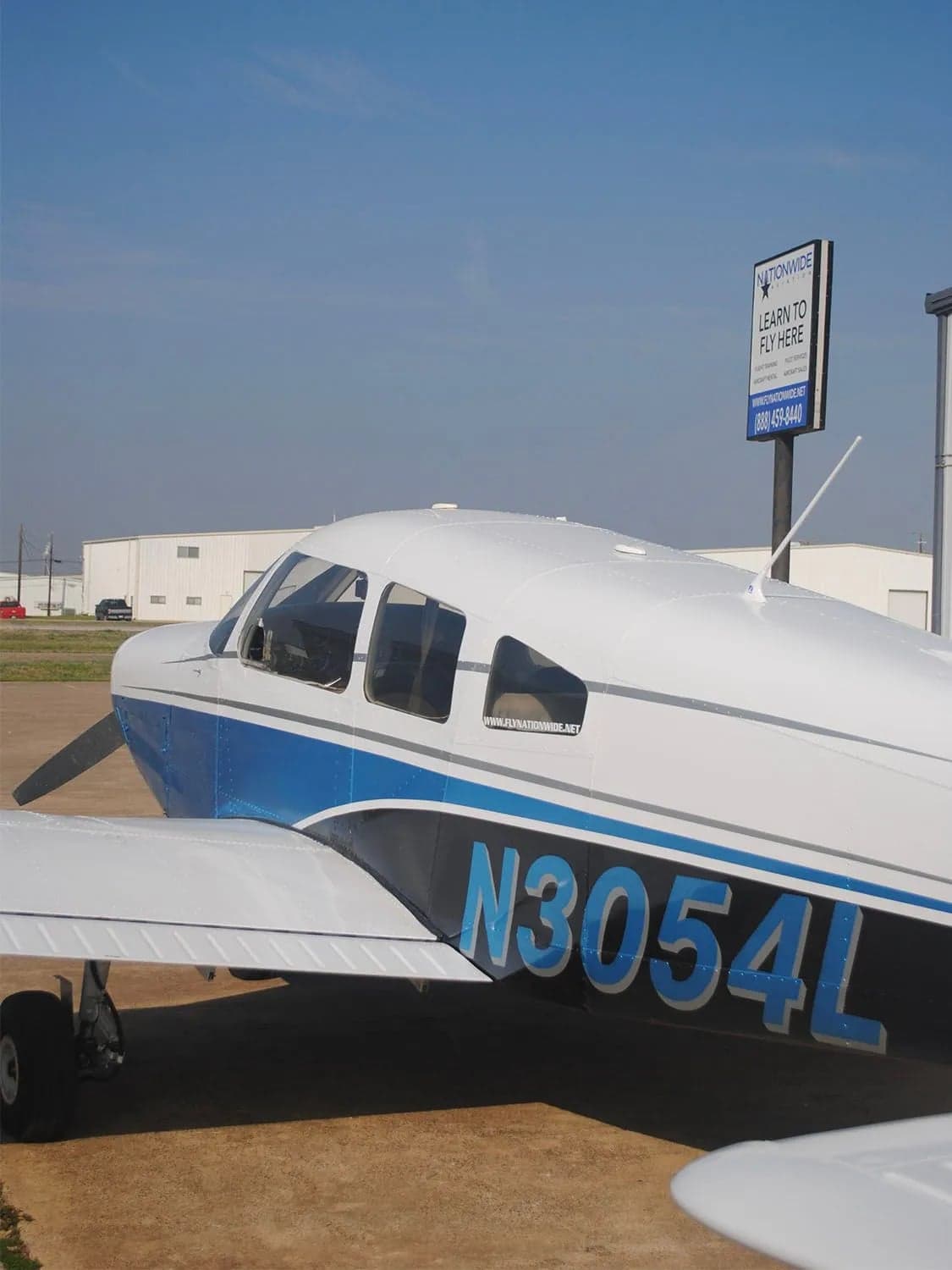 A side view of a white and blue single-engine aircraft with registration N3054L parked on a tarmac next to a "NATIONWIDE AVIATION" sign.