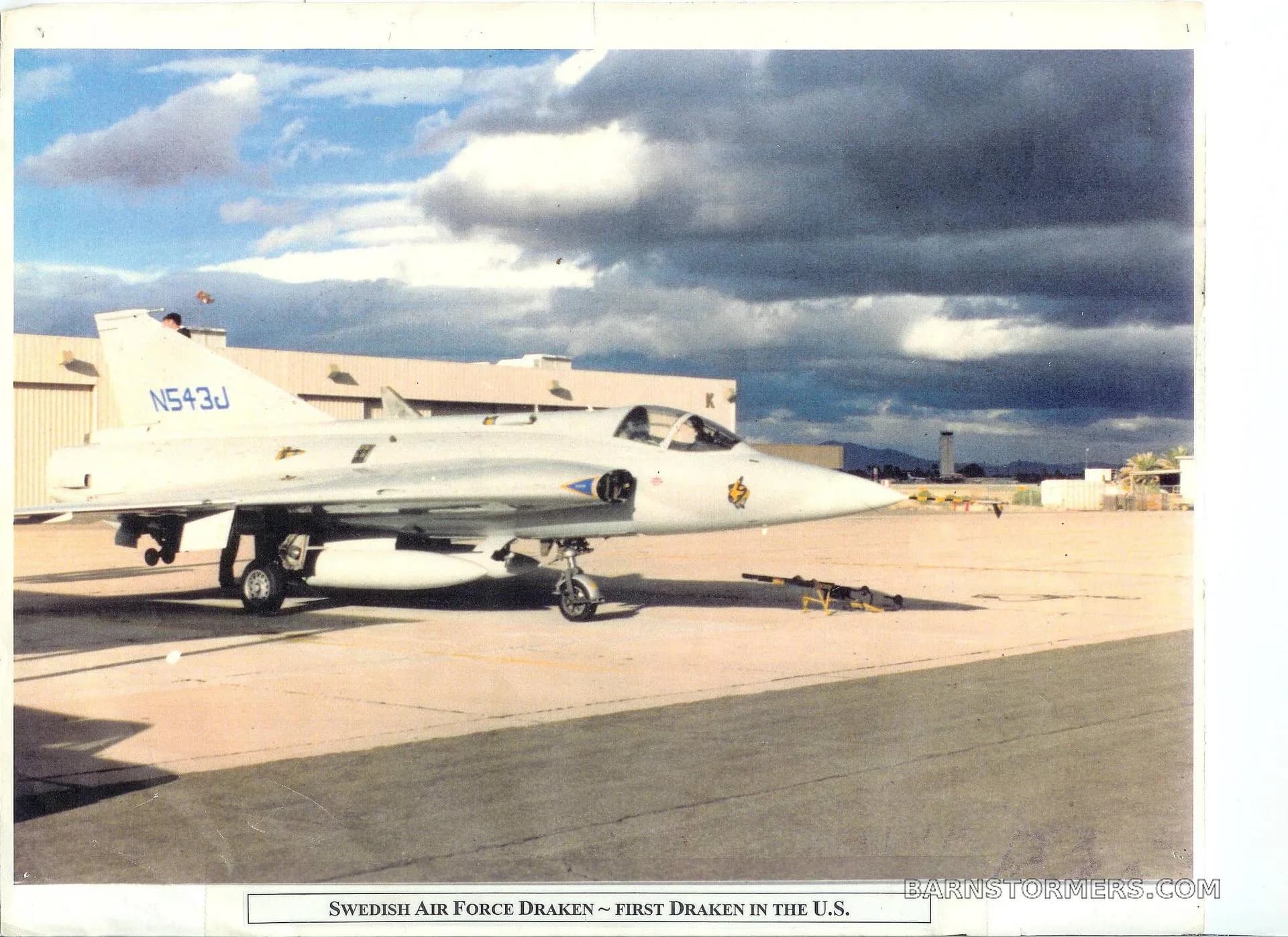 A white Saab J35 Draken aircraft with registration N543J is parked on an airfield under a cloudy sky.