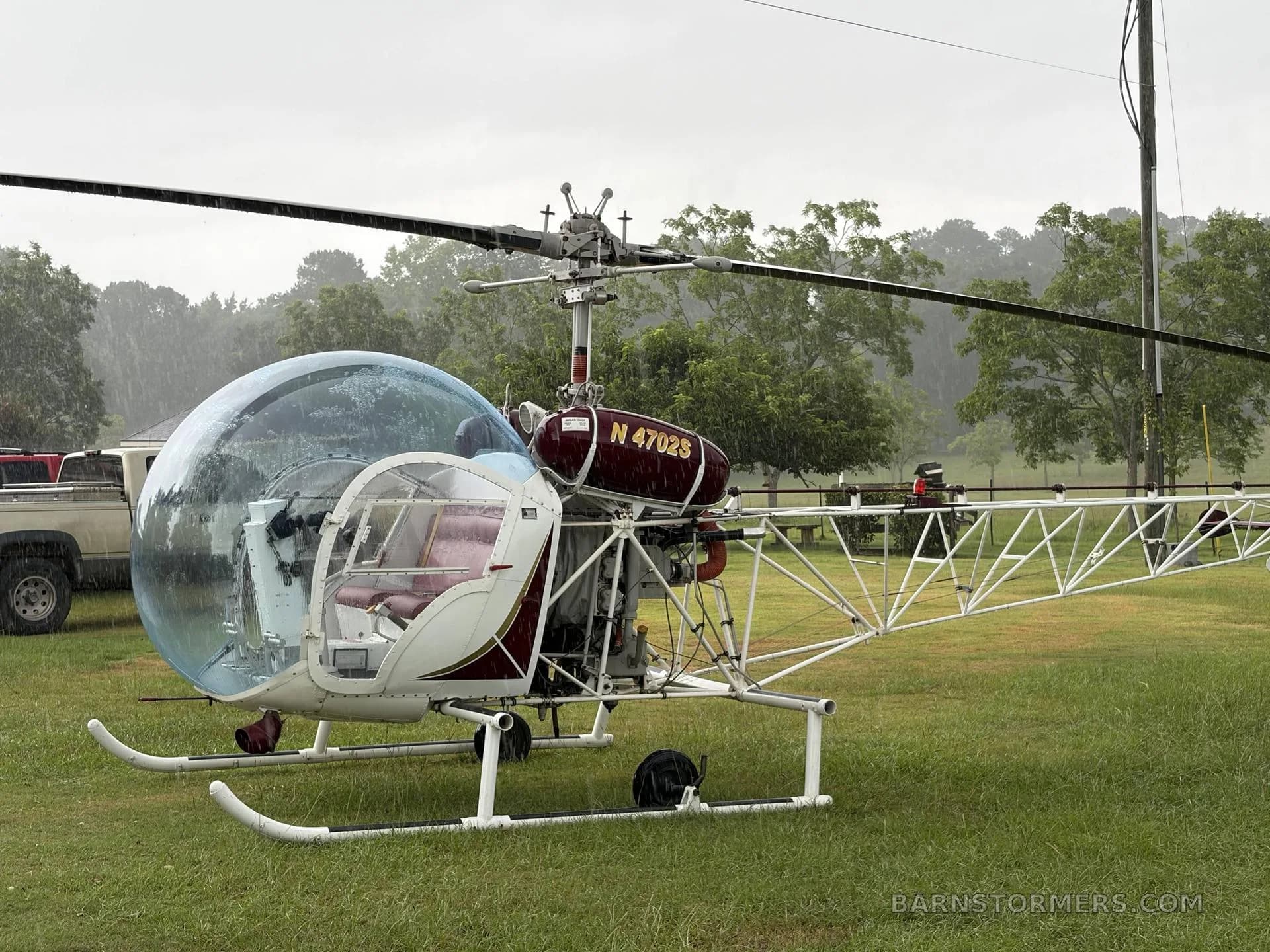 A red and white helicopter with a bubble canopy, registration N4702S, parked on a grassy field on a cloudy day.