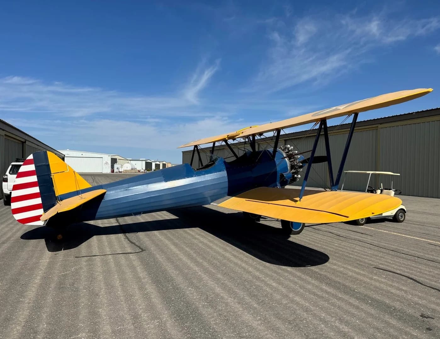 A blue and yellow biplane with a red and white striped tail parked on an asphalt tarmac next to hangars.