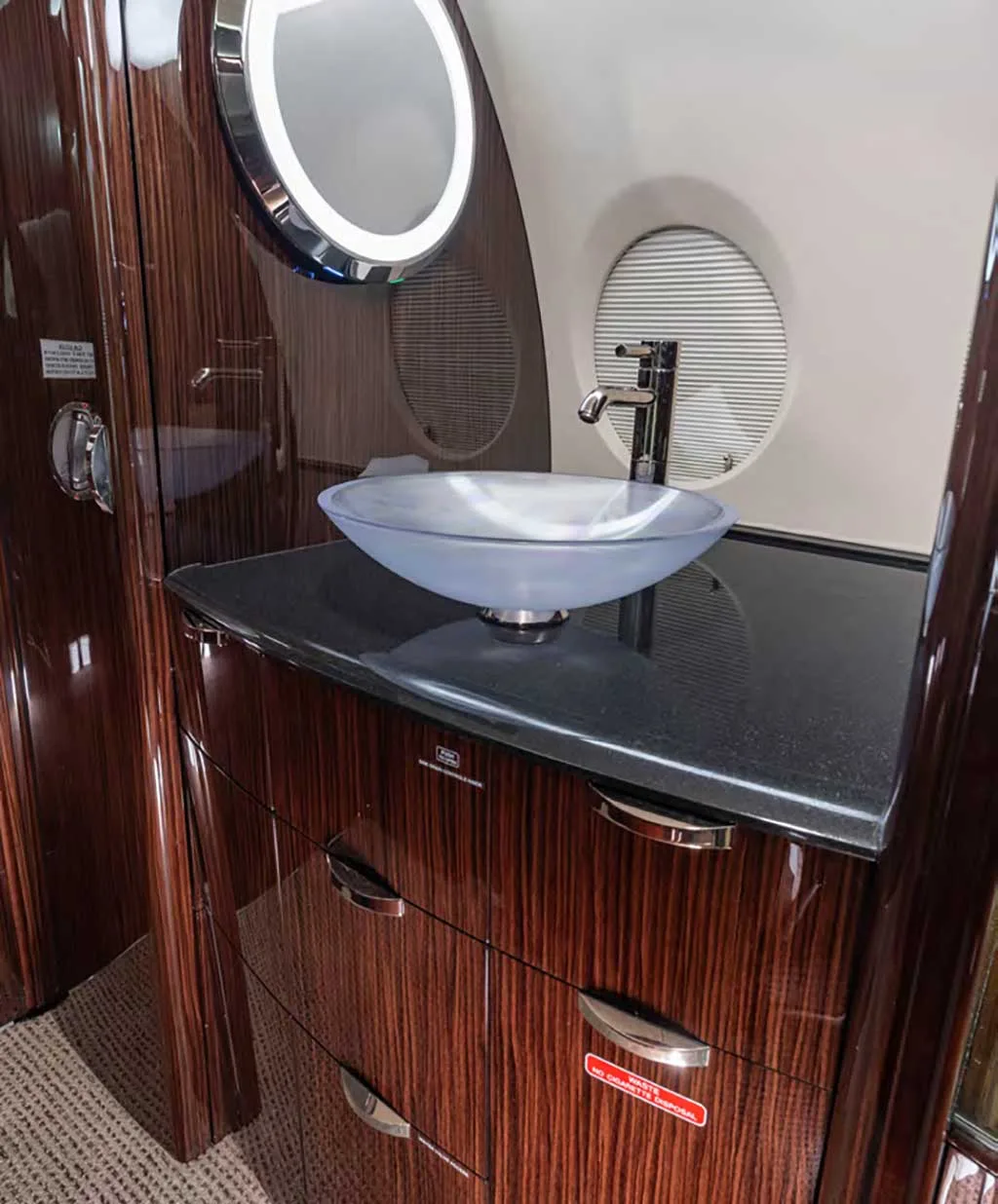 A modern lavatory sink with a clear glass bowl and dark wood vanity inside the aircraft.