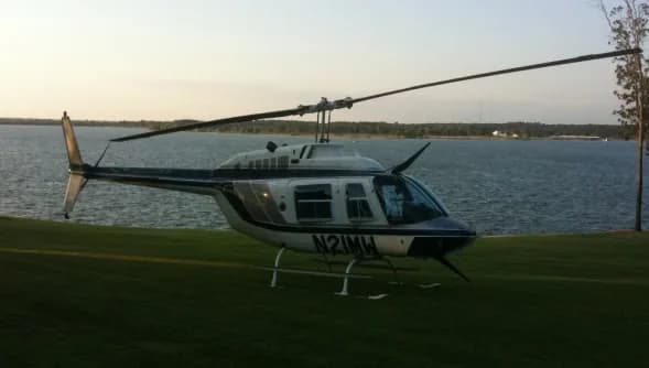 A Bell 206 Jet Ranger helicopter is parked on a grassy area near a lake.