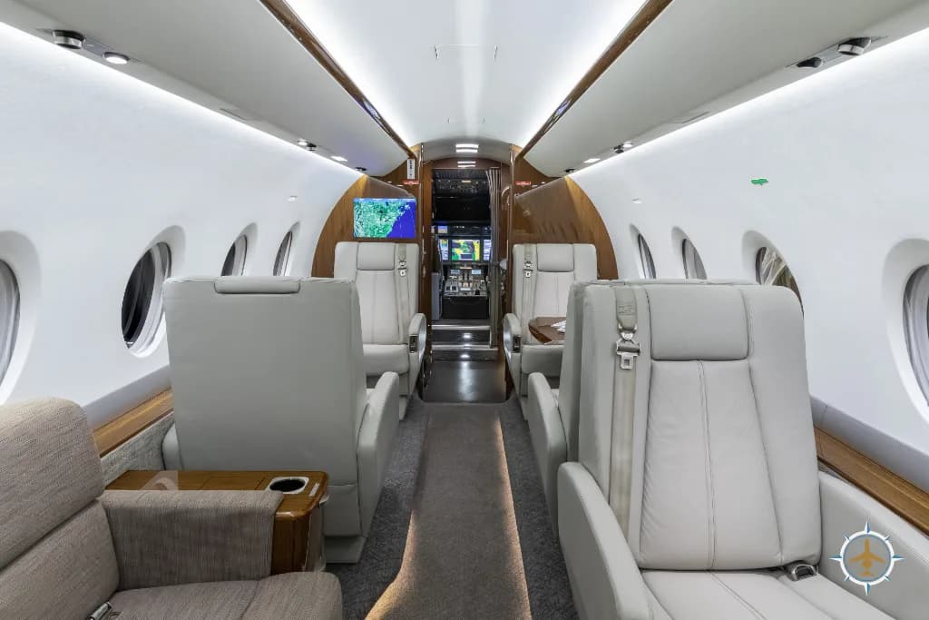The business jet cabin interior, with light grey seats and a beige sofa, looking towards the cockpit area.