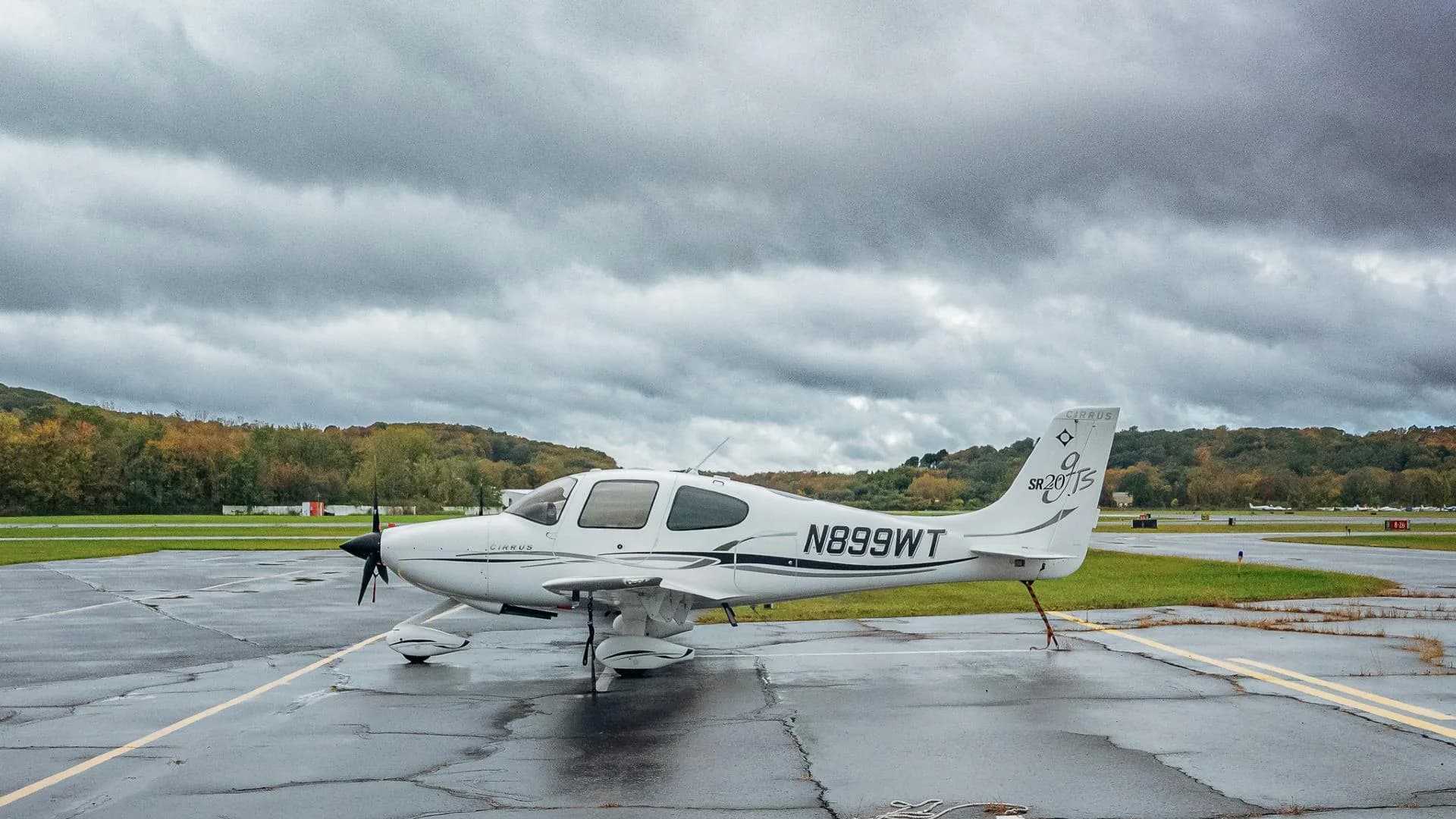 A white Cirrus SR20 aircraft with black and grey accents, registration N899WT, parked on a wet tarmac under a cloudy sky.