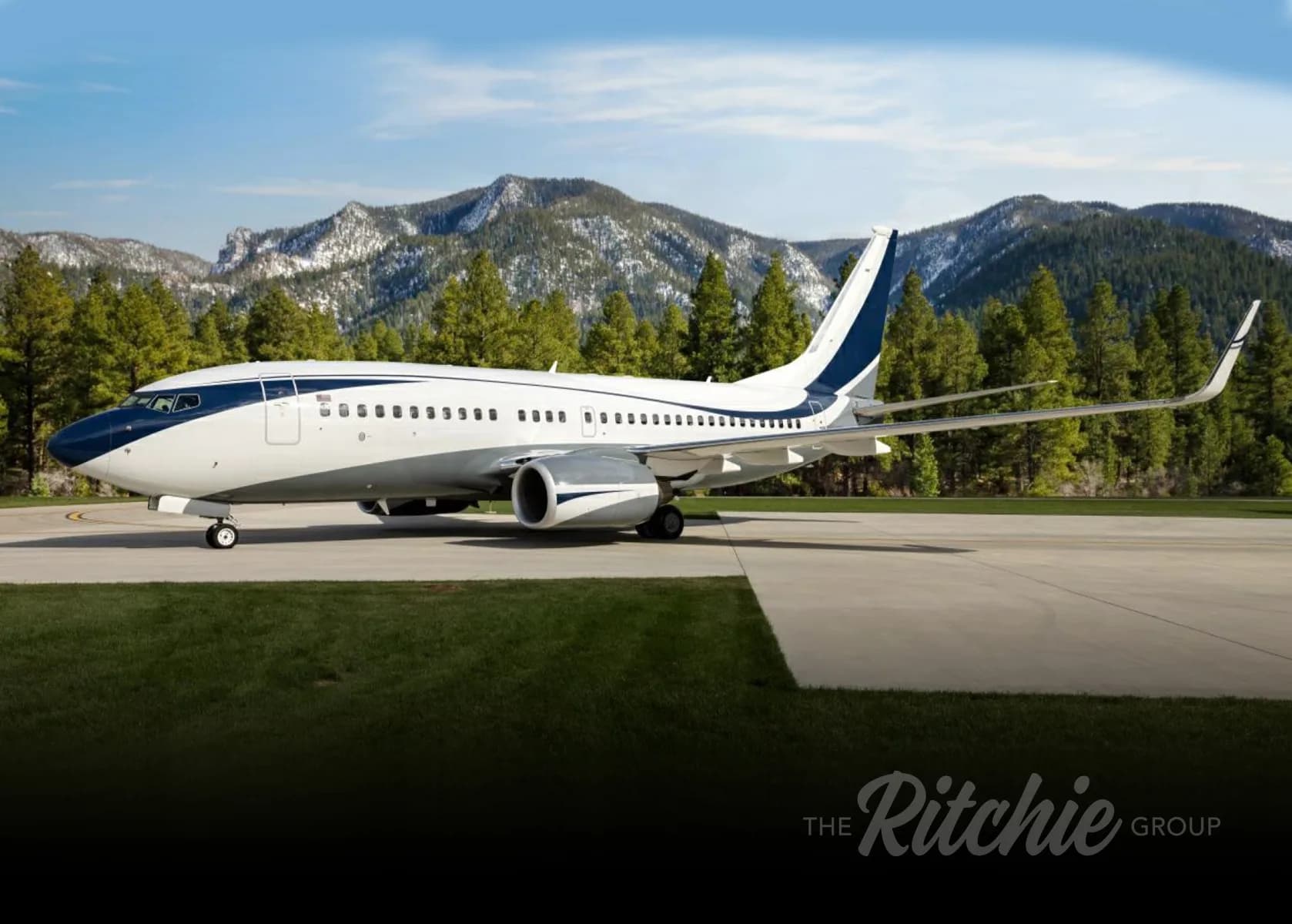 A white and dark blue Boeing 737 BBJ parked on an airfield with snow-capped mountains and pine trees in the background.