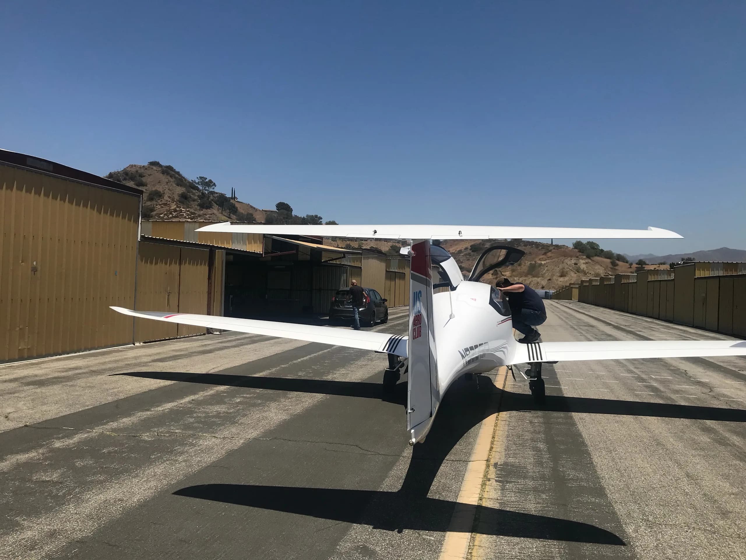 A white Pipistrel Panthera aircraft with red and gray accents, viewed from the rear, on an airfield with hangars.