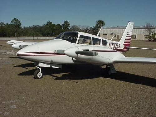 A Piper Apache aircraft with a white and maroon paint scheme is parked on the grass.