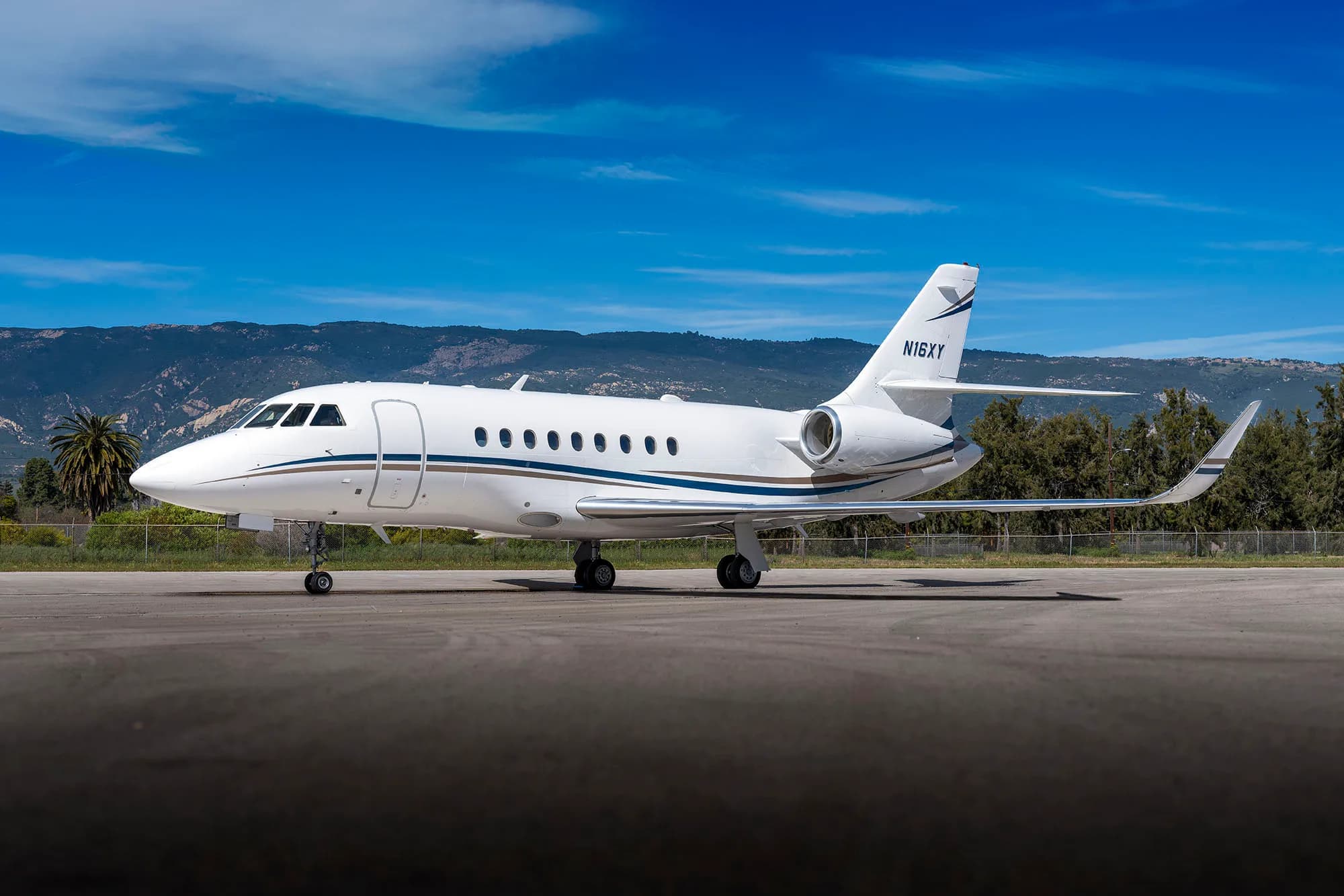 A white Falcon aircraft with blue and gold stripes sits on the tarmac with trees and mountains in the background.