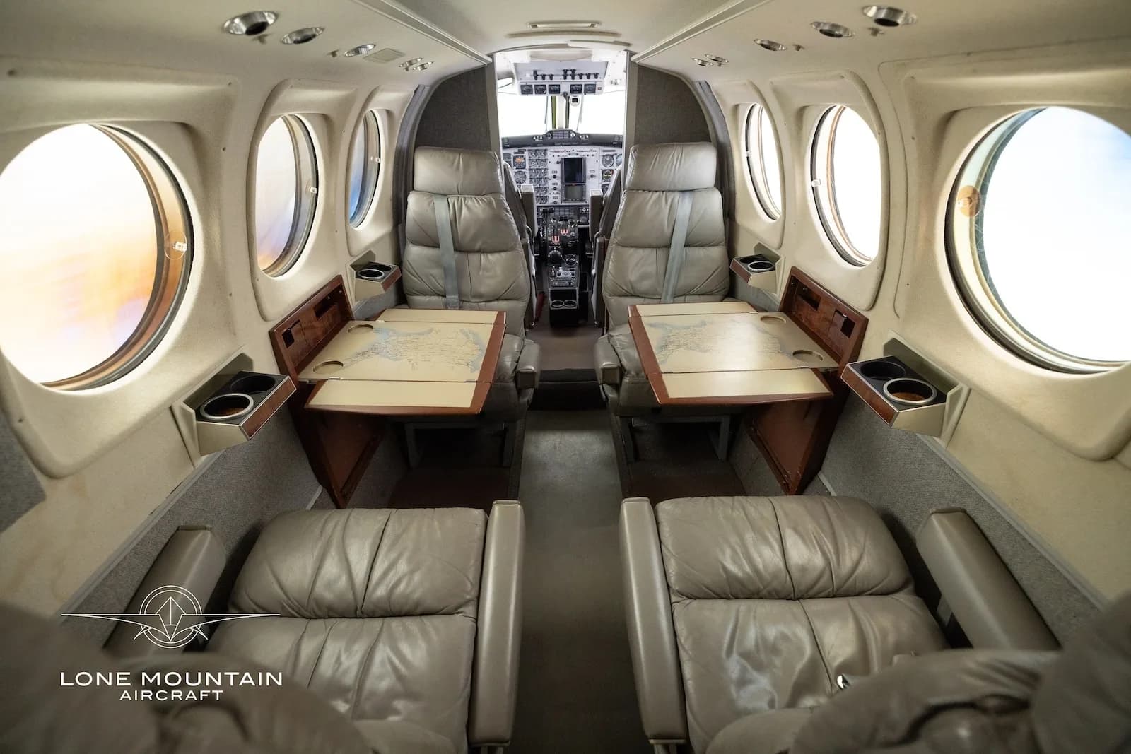 The aircraft's interior cabin features grey leather seats and two tables with maps, looking towards the cockpit.