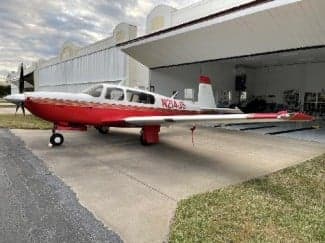 A red and white Mooney M20C aircraft, registration N214JS, parked outside a hangar.