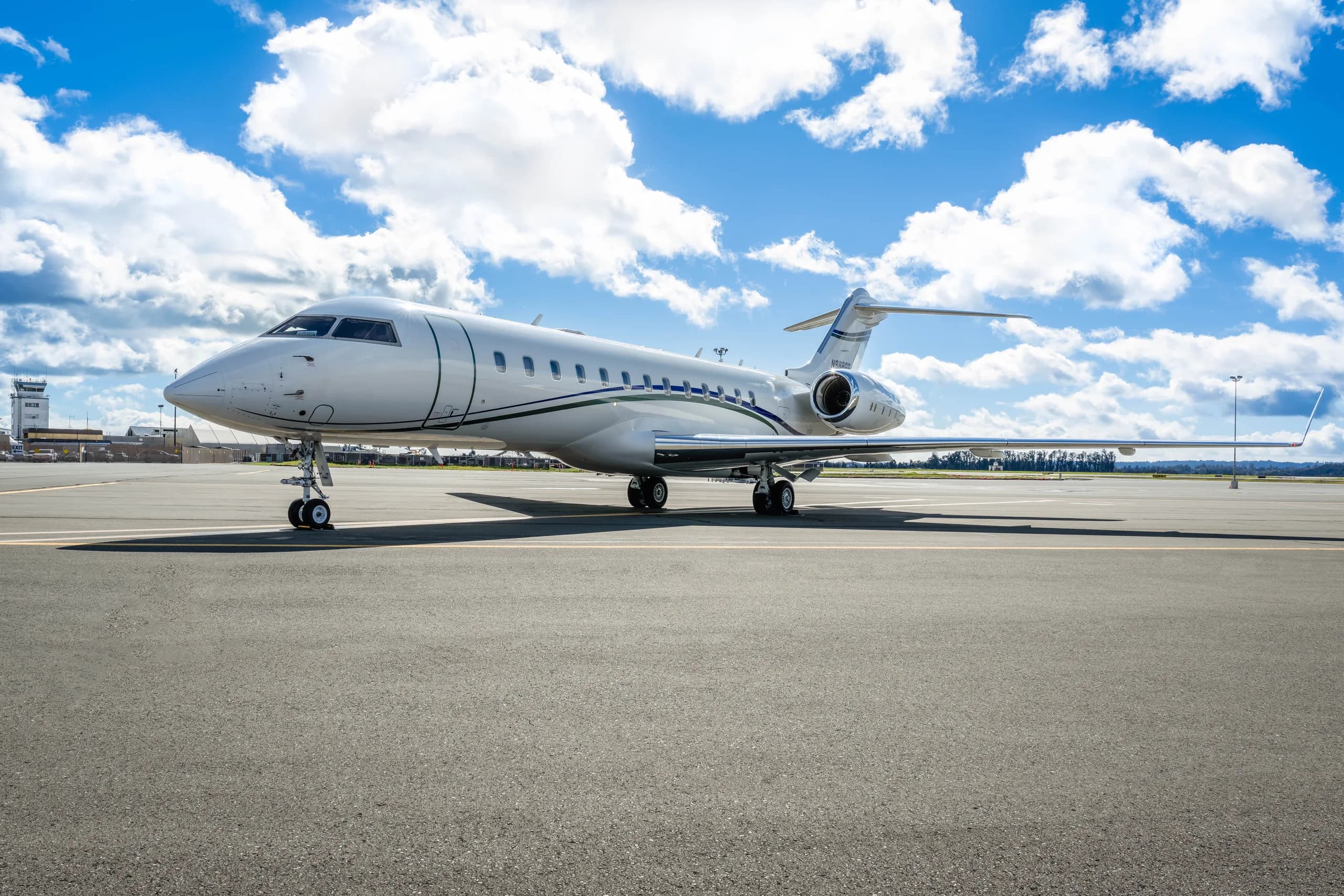 A white Bombardier Global 6000 business jet with blue and green stripes on the fuselage and tail, parked on an airport tarmac under a blue sky with clouds.