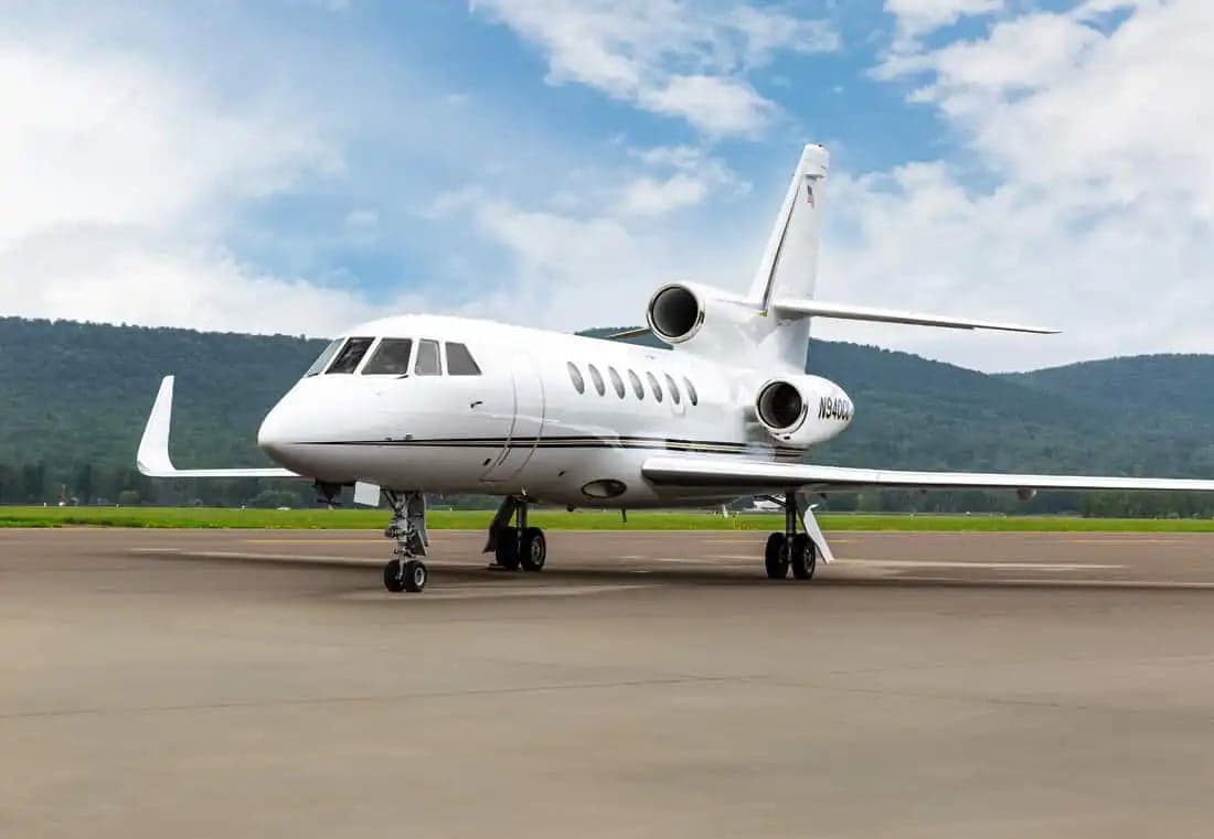 A Falcon jet sits on the tarmac with mountains in the background.