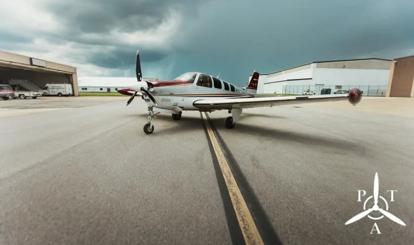 A white and red single-engine propeller aircraft, a Beechcraft Bonanza, parked on an asphalt runway under a dramatic cloudy sky.
