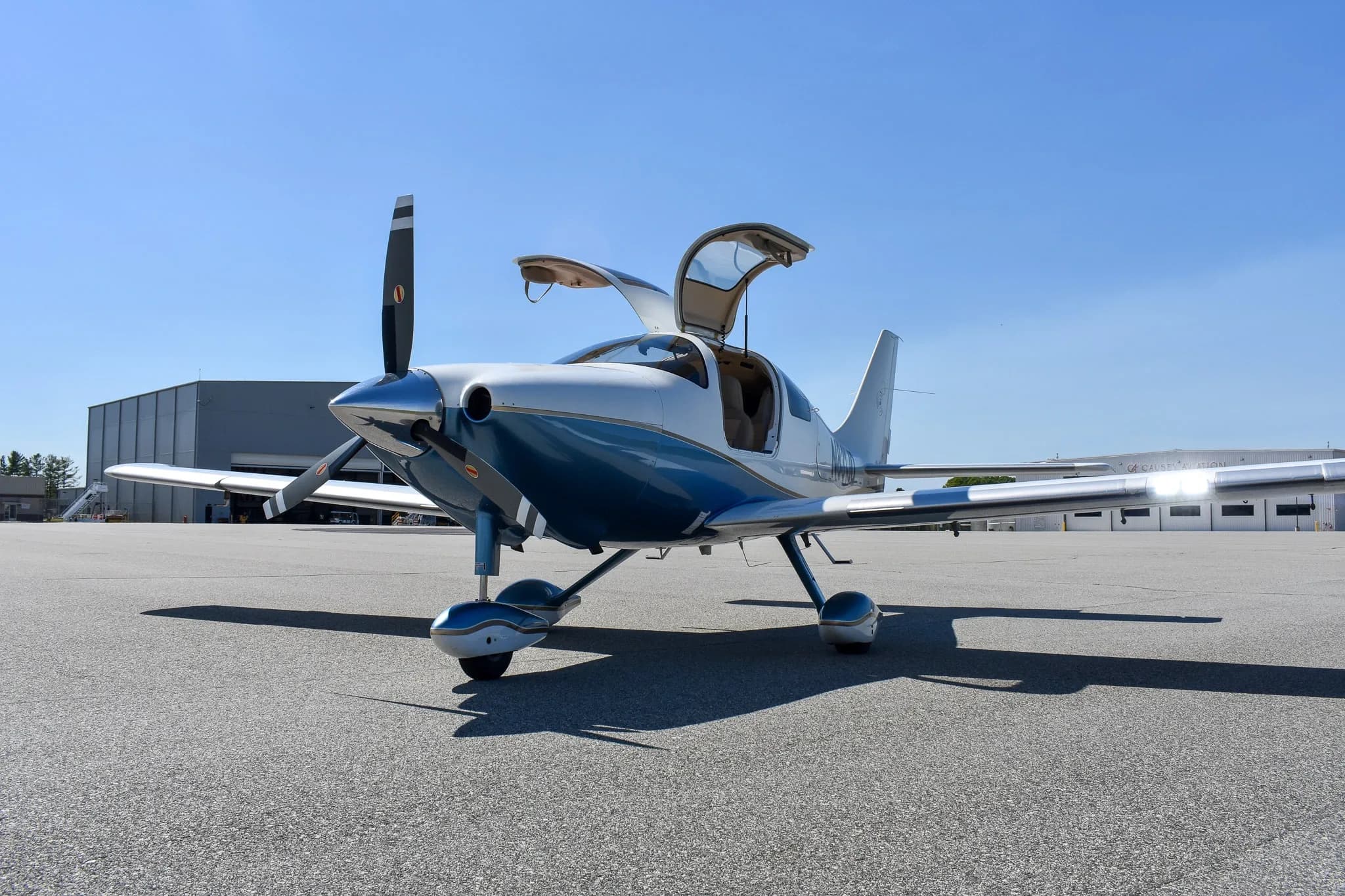 Wide front view of a white and blue single-engine aircraft with open gull-wing doors on an airfield with a hangar in the background.