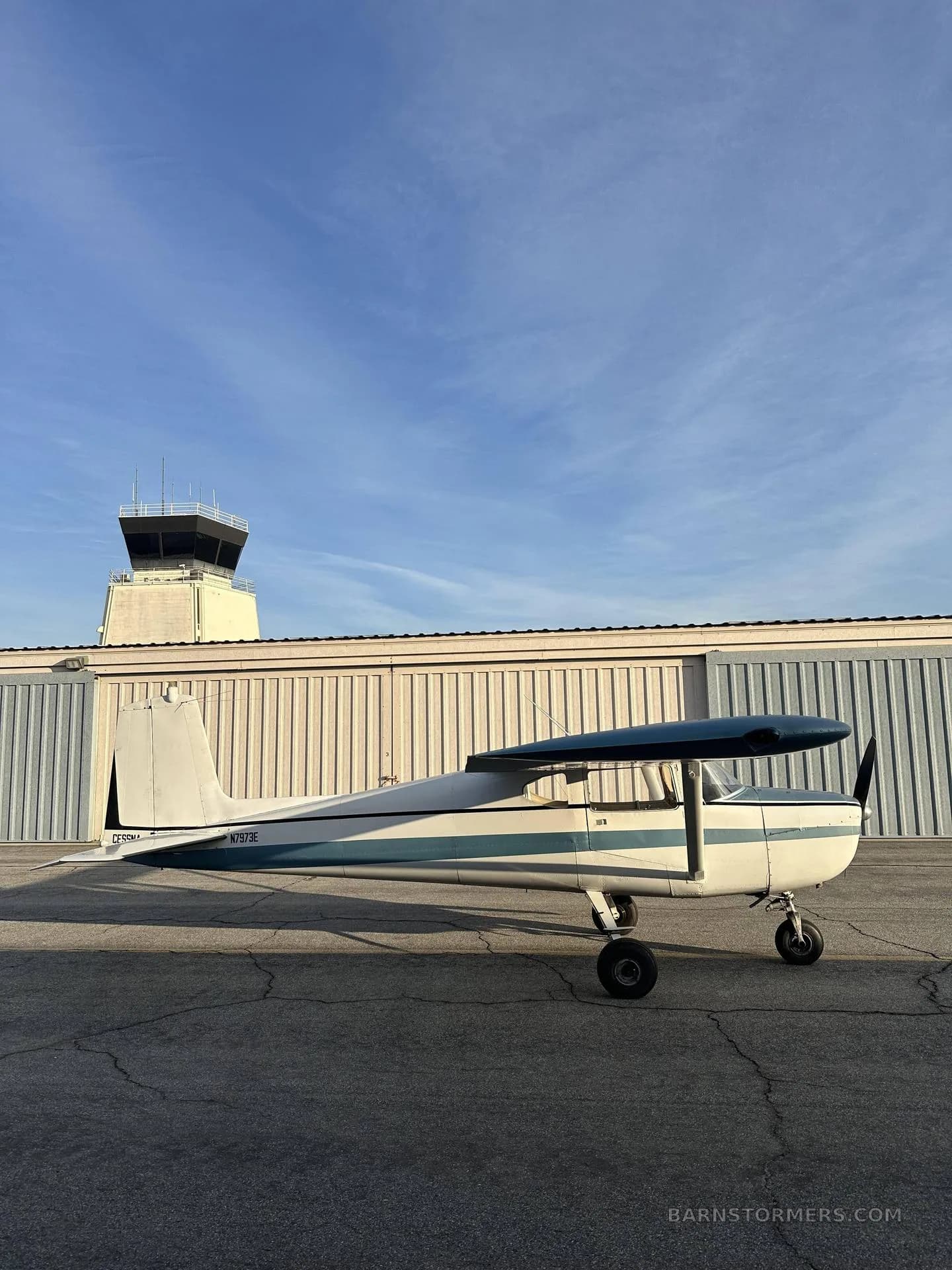 A side profile of a white and blue Cessna 150/152 aircraft parked on a tarmac with an air traffic control tower in the background.