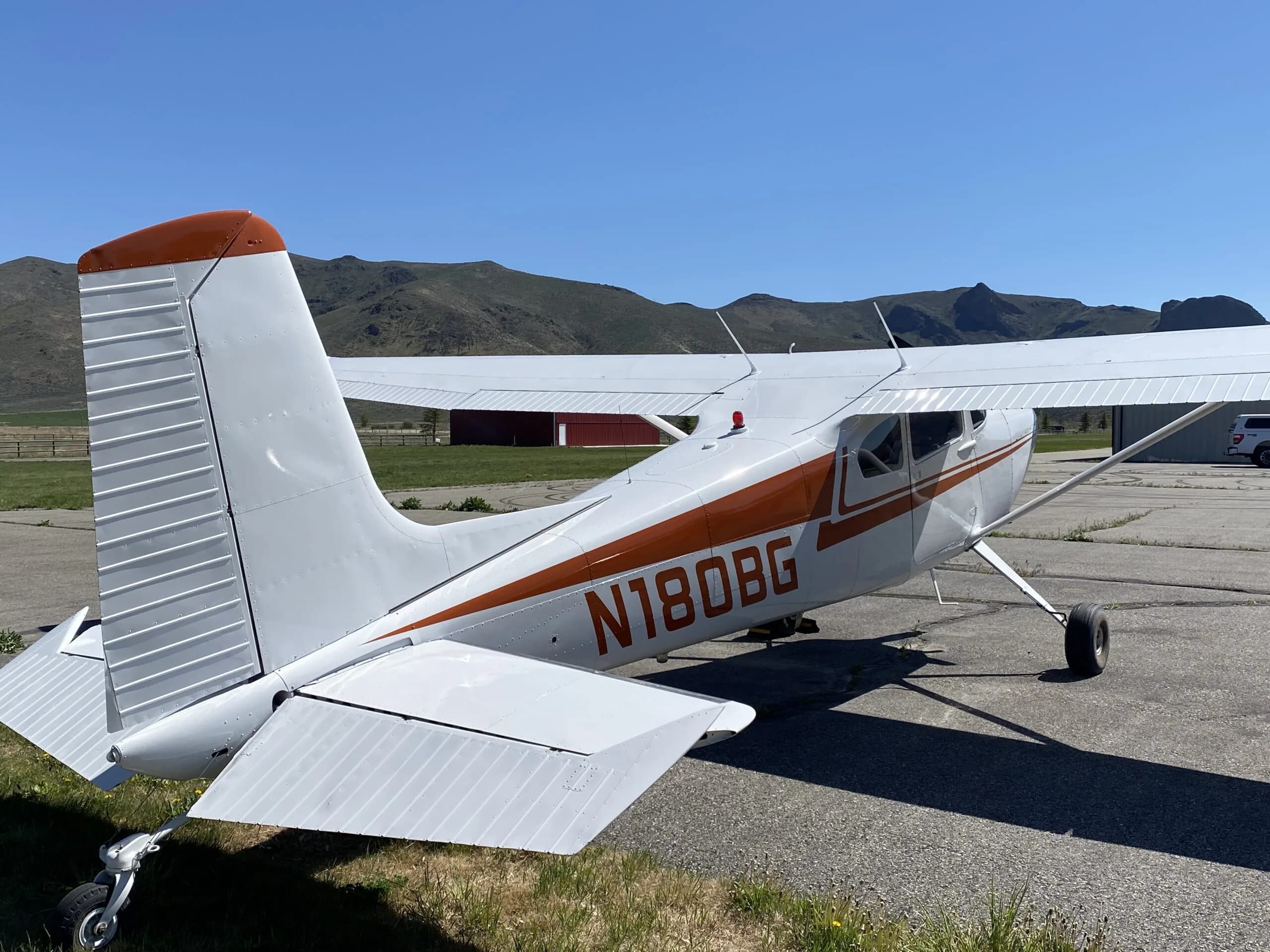 Rear view of a white Cessna 170B with orange stripes and N180BG registration parked on an airfield.