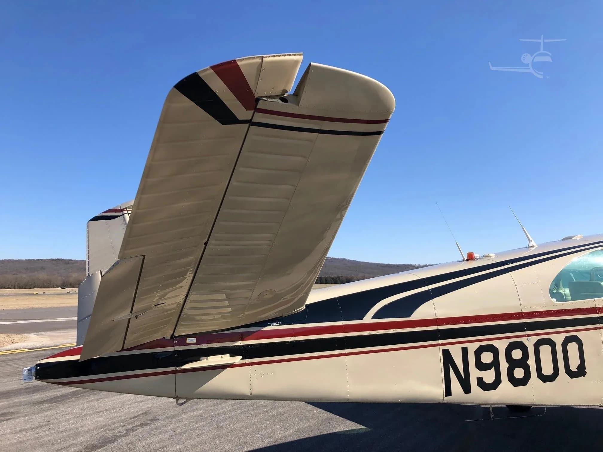 A close-up view of the V-tail and rear fuselage of the beige Beechcraft Bonanza N980Q, showing the red and black stripes.