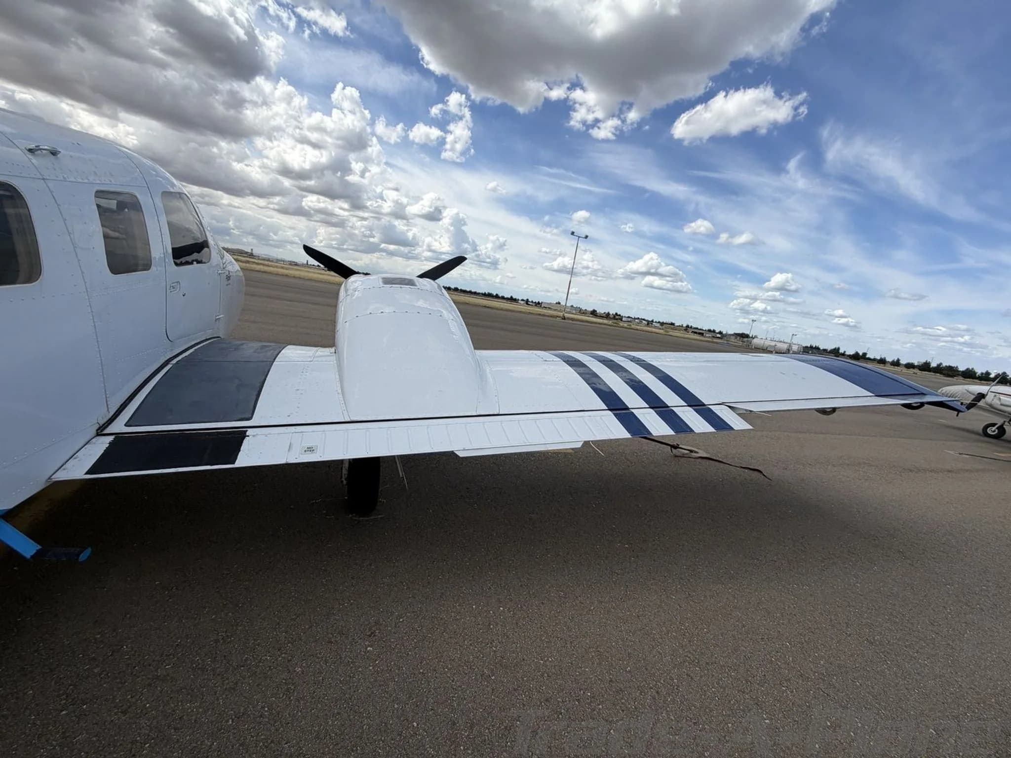 A close-up view of the left wing of the white Piper Seneca II aircraft with blue stripes, showing the engine nacelle, on an airport tarmac.