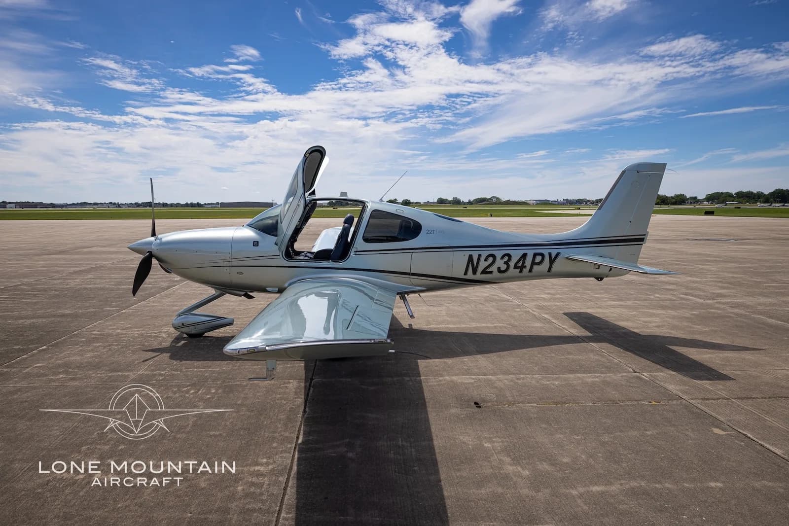 A silver Cirrus SR22 aircraft (N234PY) with its doors open, parked on a tarmac with a blue sky and clouds.